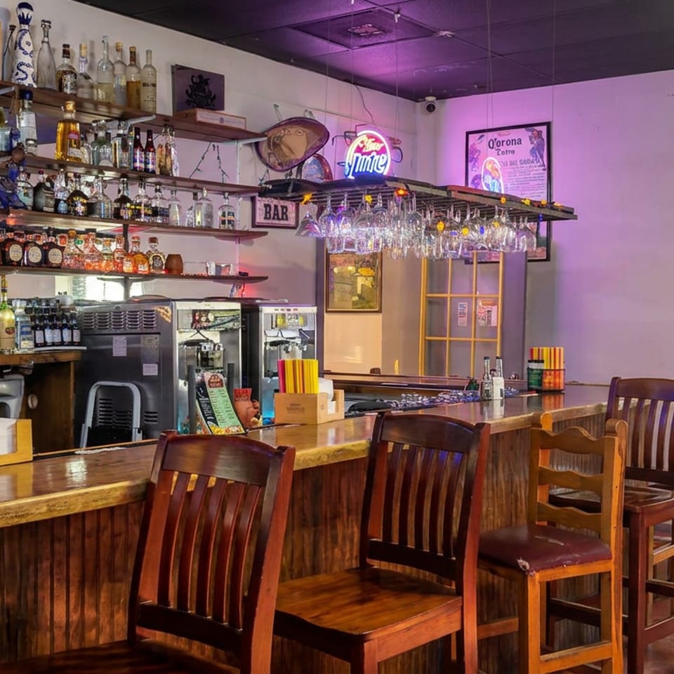 Empty restaurant bar with wooden counters, liquor shelves, hanging glassware, high chairs, and purple ambient lighting.
