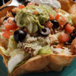 tortilla with the form of a bowl inside beans tomatoes lettuce pico de gallo back olives guacamole